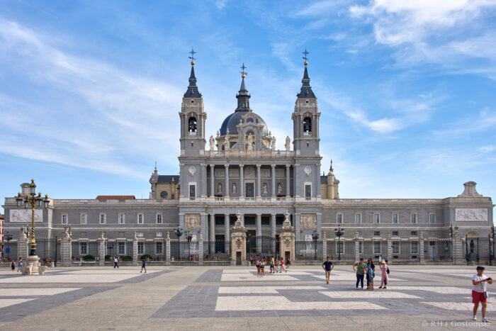 The image shows the grand façade of Almudena Cathedral in Madrid, Spain, under a blue sky. People are walking and gathering in the spacious square in front of the cathedral.