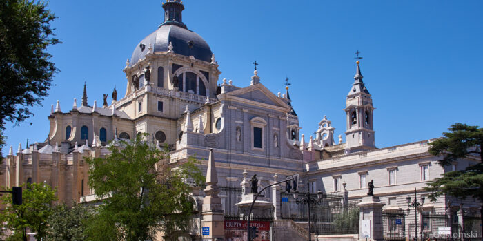 Almudena Cathedral, Madrid A large stone cathedral with a domed roof, towers, and intricate architectural details stands against a clear blue sky, surrounded by trees and a black iron fence.