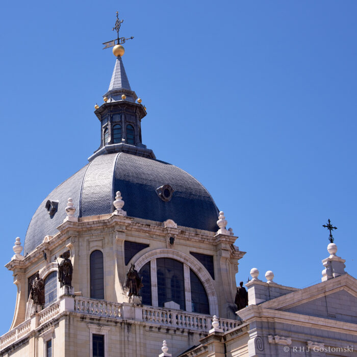 Almudena Cathedral, Madrid The image shows the dome of a grand, historic building with statues along the balustrade, topped with a weathercock against a clear blue sky.