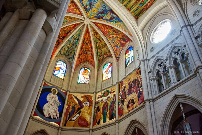 Almudena Cathedral, Madrid Ornate cathedral interior with colourful painted vaulted ceiling, stained glass windows, and detailed religious frescoes depicting saints and biblical scenes on the stone walls. Light streams through arched windows.