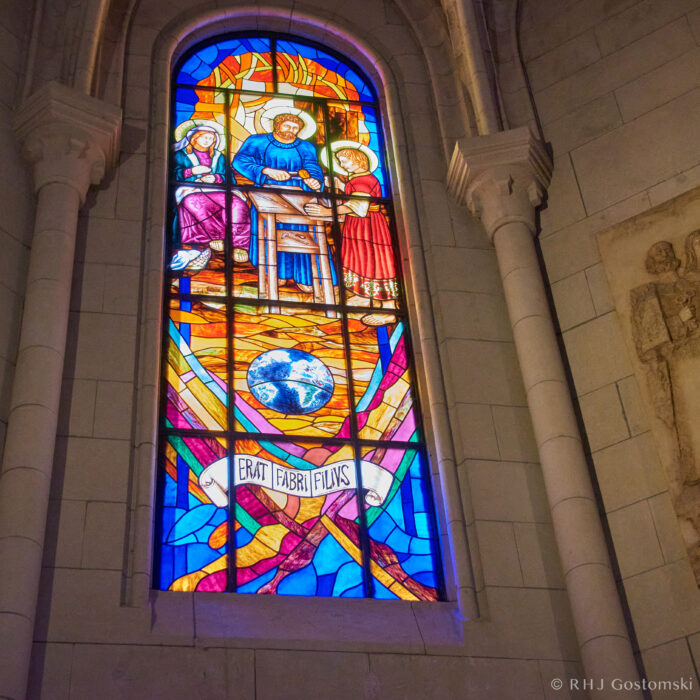 Stained glass window A colourful stained glass window shows three figures, including a man working at a table, with a globe and banner below reading ERATI FABRI FILIUS. Light shines through the vibrant blue, orange, and purple glass.