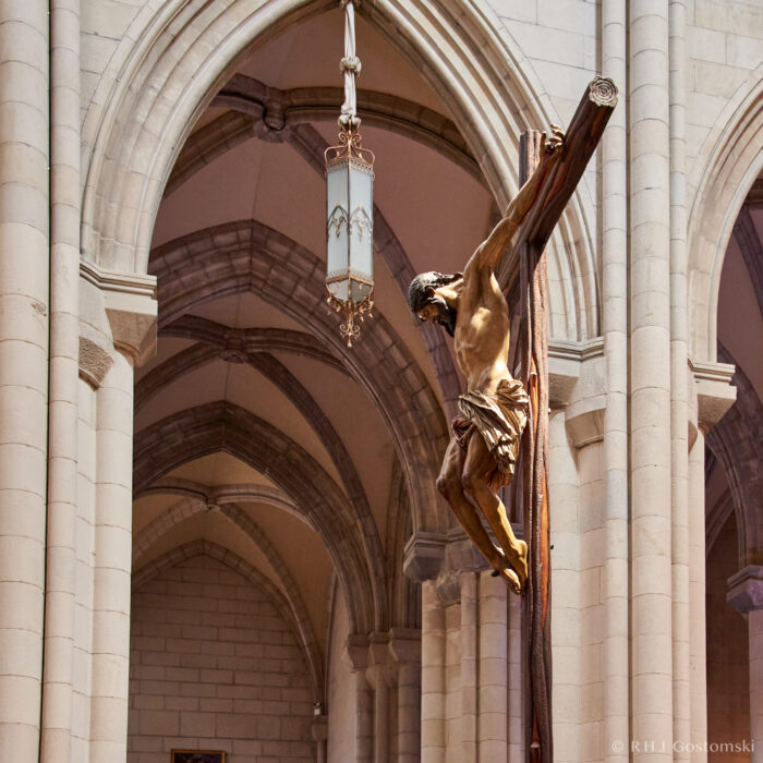 A wooden crucifix of Jesus hangs prominently in Madrid’s Almudena Cathedral, surrounded by tall arches and an ornate hanging lamp. Warm sunlight highlights the figure and the architectural details.