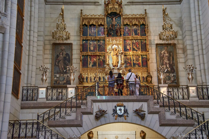 Almudena Cathedral, Madrid Three people stand on a staircase inside a grand cathedral, facing an ornate golden altar with religious artwork and statues. The space is adorned with chandeliers and decorative details.
