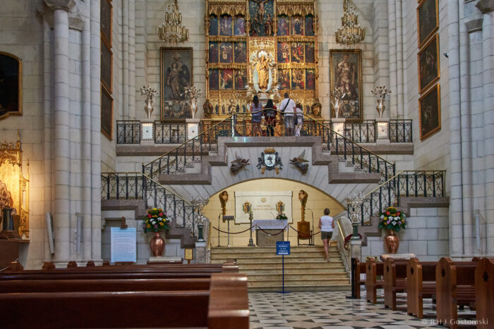 Almudena Cathedral, Madrid A grand church interior with wooden pews, ornate altar, gilded religious artwork, and several people admiring the artwork at the front near a staircase and decorative railings.