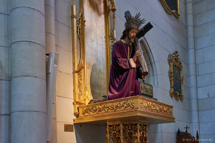 Almudena Cathedral, Madrid A statue of Jesus wearing a purple robe and crown of thorns stands on an ornate golden platform inside a church, with a cross behind him and decorative elements on the surrounding walls.