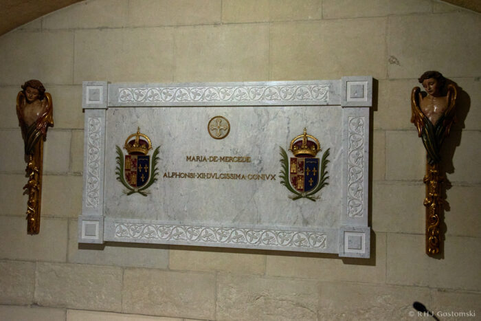 Almudena Cathedral, Madrid A marble plaque on a stone wall, bordered with ornate carvings and flanked by two angel sculptures. The plaque bears royal crests and inscriptions in Latin, honouring Maria de Mercede and Alphonsi.