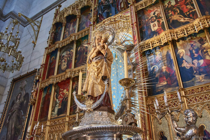 Almudena Cathedral, Madrid A golden statue of the Virgin Mary holding baby Jesus stands before an ornate altarpiece adorned with religious paintings, gold details, and candelabra in a richly decorated church interior.
