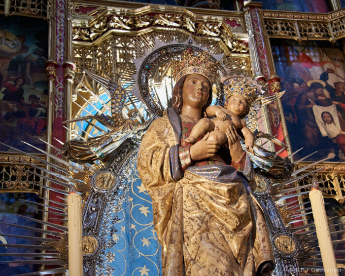 Almudena Cathedral, Madrid A richly adorned statue of the Virgin Mary holding baby Jesus, both wearing ornate crowns and robes, stands in front of a gold and blue altar decorated with religious paintings and radiating golden beams.