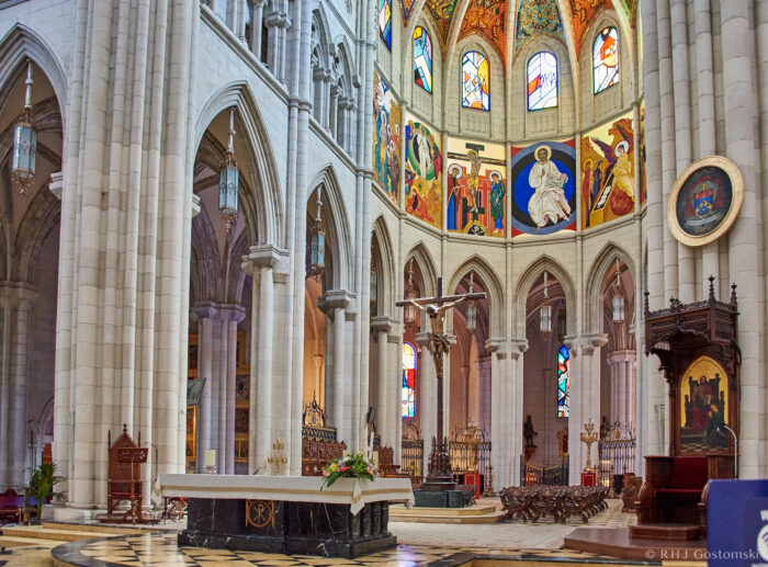 Almudena Cathedral, Madrid Interior of a large, ornate cathedral with tall stone arches, colourful stained glass windows, vibrant religious paintings, and an altar at the centre beneath a vaulted dome. Several chairs and intricate decorations are visible.