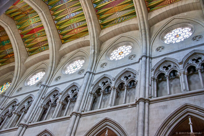Interior view of Almudena Cathedral in Madrid, featuring tall stone arches, stained glass rose windows, and a colourful, patterned vaulted ceiling above.