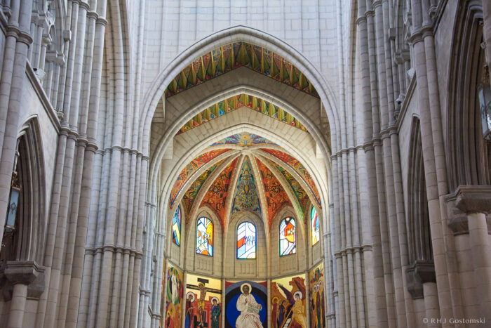 Almudena Cathedral, Madrid Stained glass windows and colourful ceiling murals decorate the interior of a grand cathedral with tall, arched stone columns and intricate architectural details.