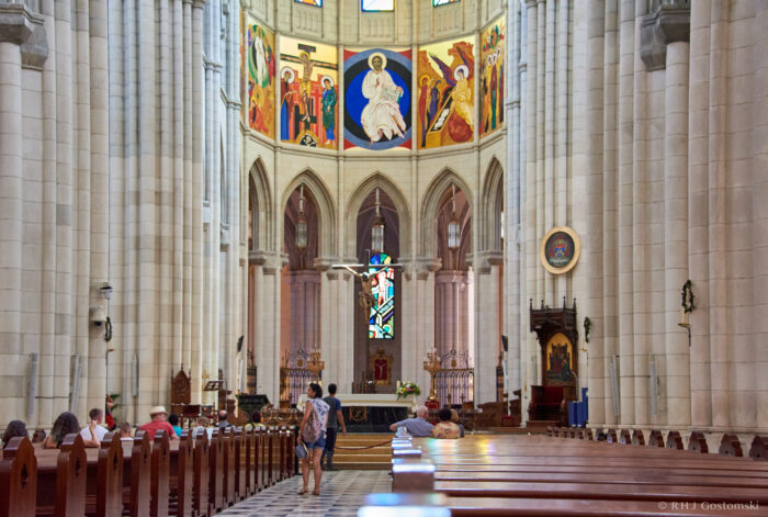 Interior of the Almudena Cathedral, Madrid, with high vaulted ceilings, stained glass windows, colourful religious paintings above the altar, and several people seated or walking among wooden pews.