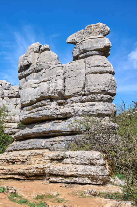 Torcal-NZ6_5855 Large, weathered limestone rock formation resembling the profile of a human face against a bright blue sky, surrounded by sparse vegetation and rocky ground.