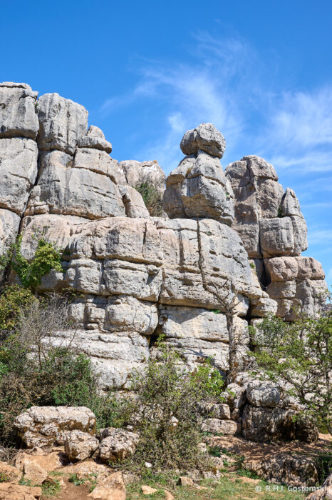 Torcal-NZ6_5872 Large, weathered limestone rock formations rise against a bright blue sky with wispy clouds, surrounded by shrubs and sparse vegetation at the base.