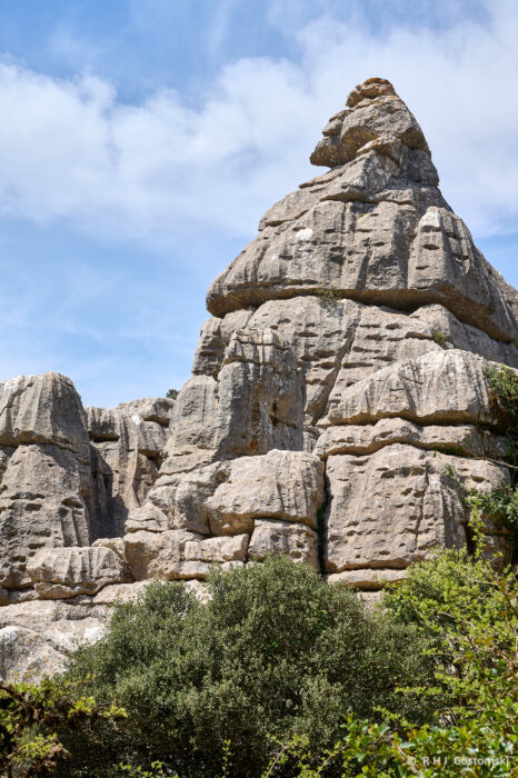 Torcal-NZ6_5913 Large, rugged limestone rock formation with layered, jagged edges rises above green shrubs against a blue sky with scattered clouds.
