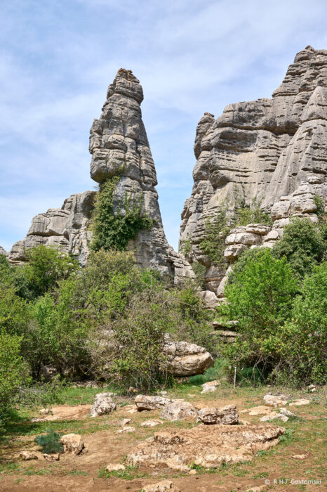 Torcal-NZ6_5936 Tall, narrow rock formation rises among rugged cliffs and green shrubs under a partly cloudy sky, with rocky ground and scattered vegetation in the foreground.