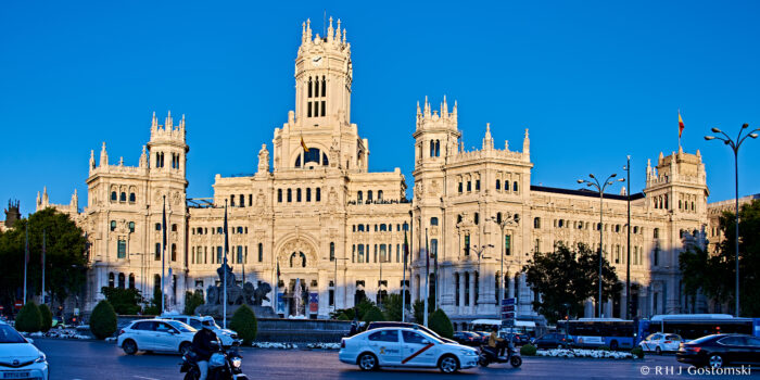 Photo by Rom Gostomski A grand, ornate white building with multiple towers, the Palacio de Cibeles in Madrid, Spain, stands under a clear blue sky; cars and motorbikes move along the busy street in front.