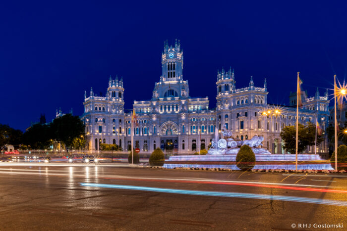 The illuminated Palacio de Cibeles and the Cibeles Fountain light up Plaza de Cibeles in Madrid at night, with car light trails across the street and a deep blue sky in the background.