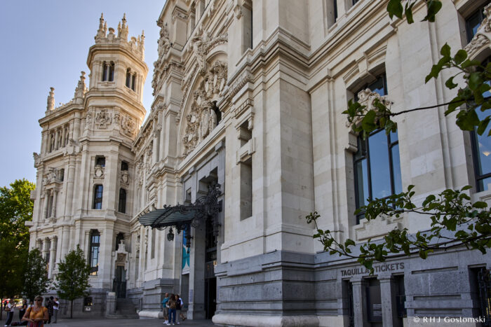 Palacio de Cibeles, Madrid A grand, ornate white stone building with tall towers and detailed carvings stands against a blue sky. People walk by its entrance, near a sign for tickets, with trees lining the foreground.