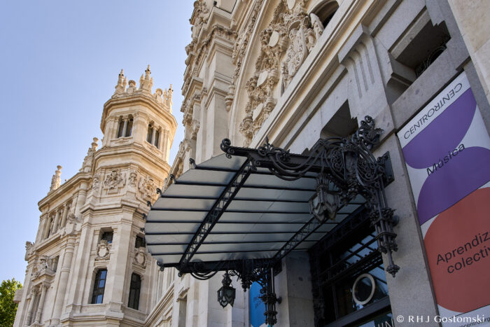 Palacio de Cibeles, Madrid Ornate façade and entrance canopy of a grand historic building with detailed stonework, next to colourful banners reading “Música” and “Aprendiz…” under a clear blue sky.