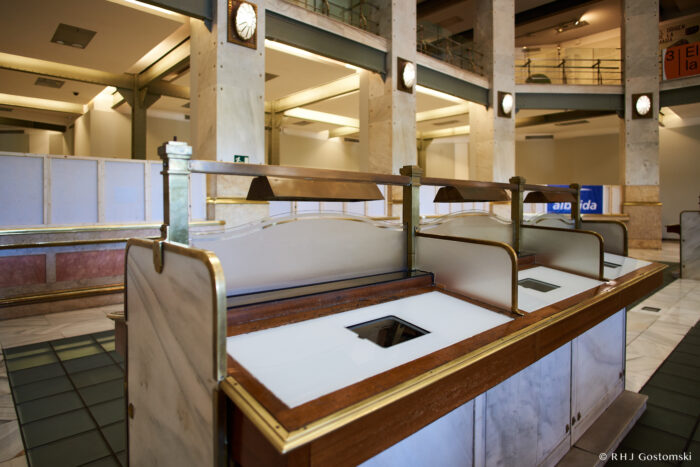 Palacio de Cibeles, Madrid A marble and wood cashier counter with glass dividers sits in a spacious, art deco-style bank lobby with tall columns, balcony railings, and two clocks mounted on pillars.