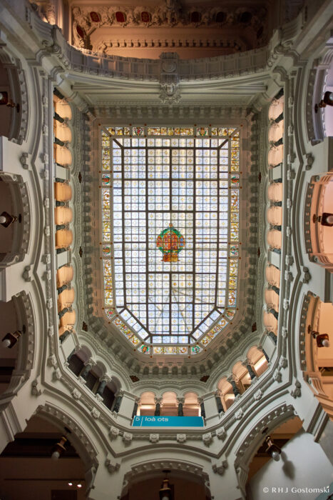 Palacio de Cibeles, Madrid Ornate glass ceiling with colourful stained-glass centrepiece, surrounded by detailed white architectural arches and columns in an elegant, historic building. A sign reading las c/s is visible near the bottom.