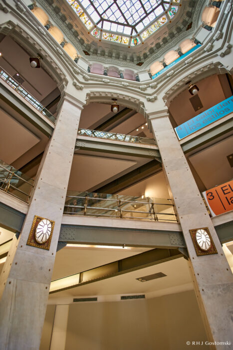 Palacio de Cibeles, Madrid Interior view of a multi-level building with a glass dome ceiling, decorative columns, and a geometric stained glass skylight. The image captures modern railings and signage on the upper floors.