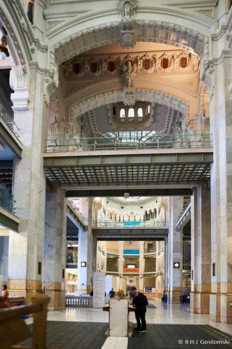 Palacio de Cibeles, Madrid Spacious grand interior of a historic building with ornate ceilings, large arched structures, marble columns, and people standing at a central information desk. Light streams in from high windows.