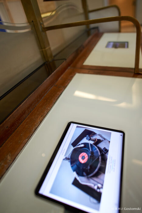 Palacio de Cibeles, Madrid A tablet displaying an image of a vinyl record on a turntable lies on a glass-topped display case with a wooden frame, in what appears to be an exhibition or museum setting.