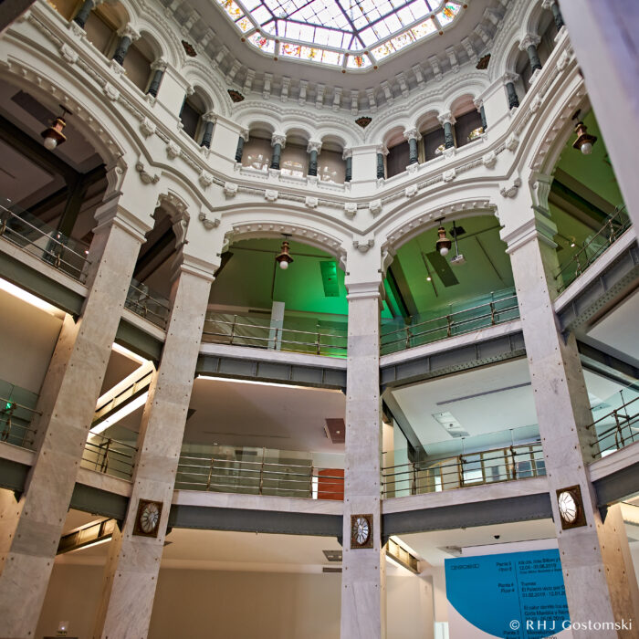 Palacio de Cibeles, Madrid A view from inside a grand, multi-storey building with marble columns, arched balconies, and a stained-glass dome ceiling. Natural light fills the spacious atrium, highlighting elegant architectural details.