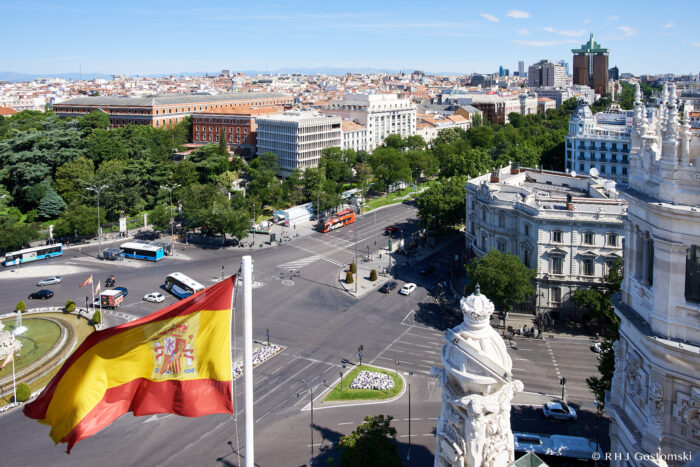 Photo by Rom Gostomski A large Spanish flag waves above Plaza de Cibeles in Madrid, with busy streets, green trees, historic buildings, and the cityscape visible under a clear blue sky.