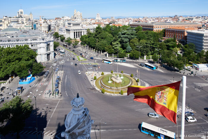 A view of Madrid from Palacio de Cibeles shows the Plaza de Cibeles with a Spanish flag in the foreground, its central fountain gleaming, and historic buildings surrounding it under a clear blue sky.