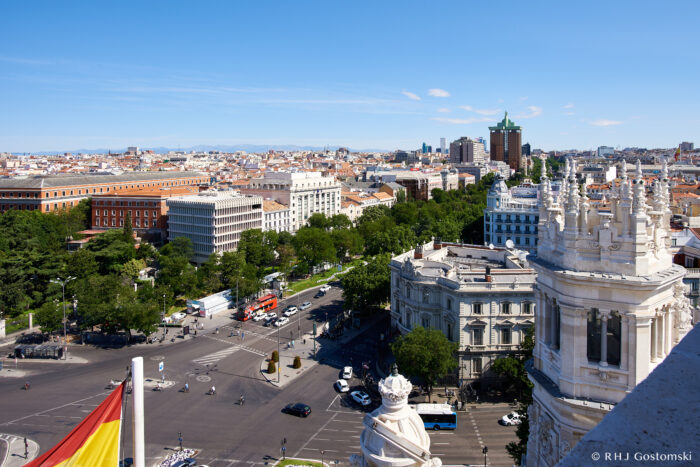 Madrid from a rooftop terrace A high view of a busy city intersection in Madrid, Spain, with historic buildings, green trees, city traffic, and the Spanish flag in the foreground under a clear blue sky.