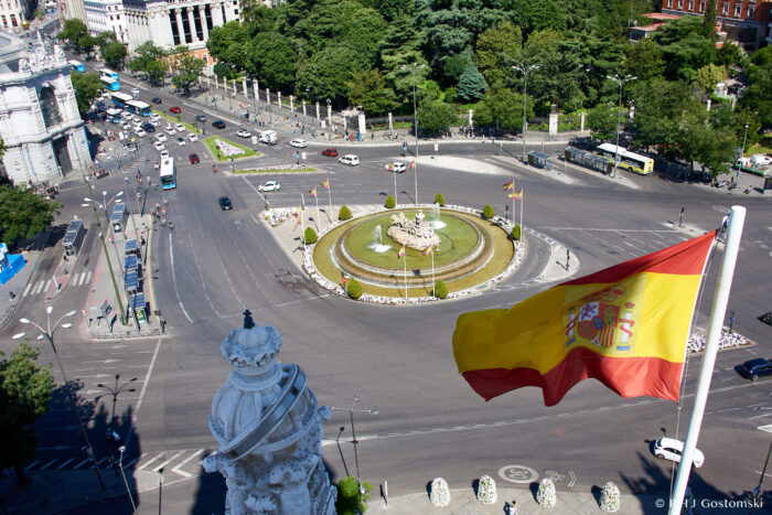Palacio de Cibeles, Madrid Aerial view of Plaza de Cibeles in Madrid, featuring a roundabout with a central fountain, surrounding greenery, traffic, and a Spanish flag prominently waving in the foreground.