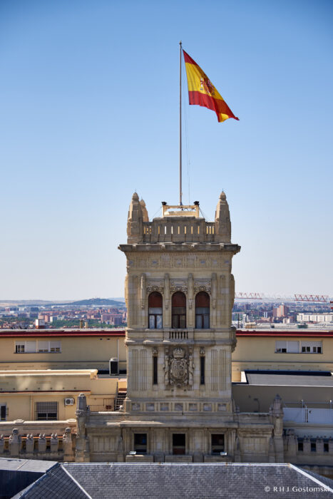 Palacio de Cibeles, Madrid A tall, ornate stone building with a large Spanish flag flying atop its central tower, set against a clear blue sky and an urban cityscape in the background.