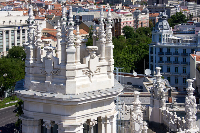 Palacio de Cibeles, Madrid Ornate white stone towers with intricate carvings overlook a cityscape featuring historical and modern buildings, trees, and red roofs under a clear blue sky.