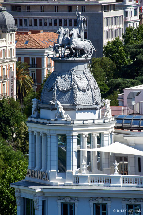 Palacio de Cibeles, Madrid Ornate rooftop of a historic white building with statues and a prominent chariot sculpture pulled by four horses, set against a backdrop of trees and city architecture. The word ABANCA is visible on the building.