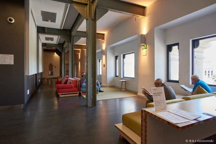 Palacio de Cibeles, Madrid A bright, modern lounge with industrial beams, large windows, and people sitting on sofas reading. A table in the foreground holds papers, and natural light fills the room.