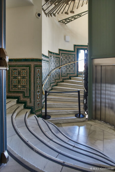 Palacio de Cibeles, Madrid A curved marble staircase with ornate, colourful tile walls ascends next to a modern lift. Light streams in from above, illuminating the steps and intricate geometric designs on the wall.