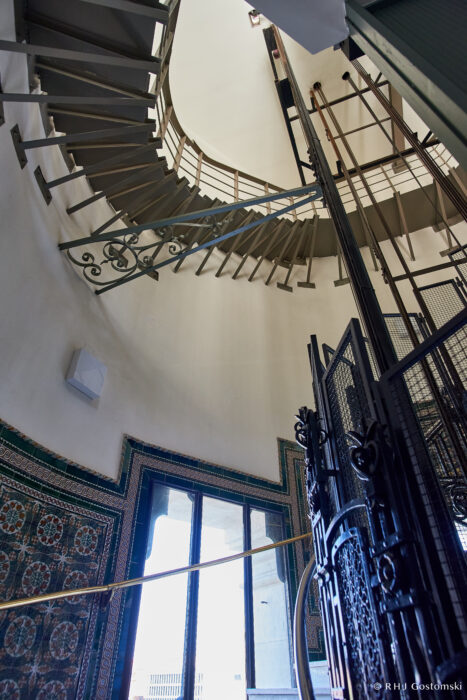 Palacio de Cibeles, Madrid A spiral staircase winds upward inside a tall, round building with patterned tiled walls, a decorative metal gate, and sunlight streaming in through large windows at the base.