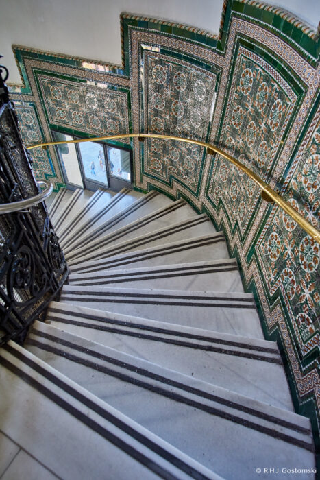 Palacio de Cibeles, Madrid A spiral staircase with white steps, intricate black railing, and walls decorated with ornate green and orange geometric tile patterns, leading down to glass doors.