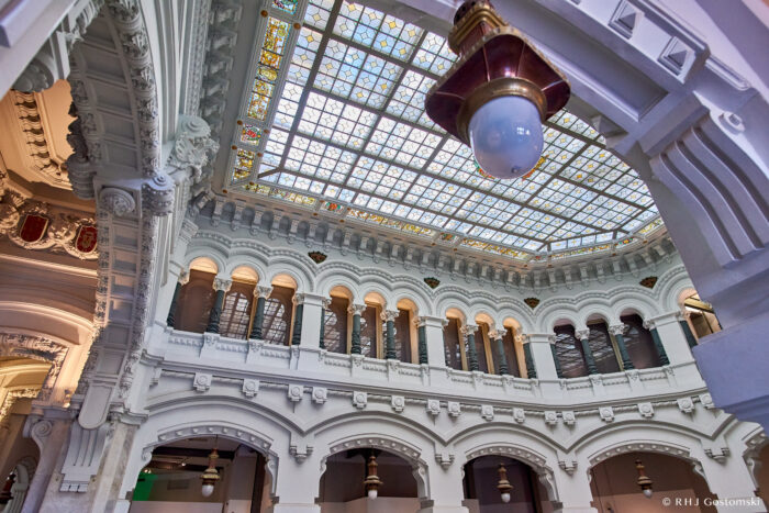 A grand interior inside Palacio de Cibeles features ornate white arches, decorative columns, and a large stained-glass skylight ceiling; an antique hanging light fitting is visible in the foreground.
