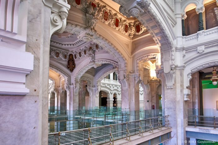 Palacio de Cibeles, Madrid A grand interior with ornate white arches, decorated ceilings with red and gold crests, and marble columns. Glass balustrades line the walkways, and soft natural light fills the spacious, elegant hall.