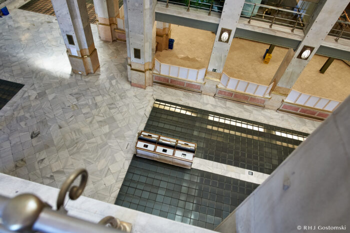 Palacio de Cibeles, Madrid View from above of a spacious, modern interior with marble and dark tiled flooring, tall columns, and a display case in the centre. Partially covered upper floors are visible, with railing in the foreground.