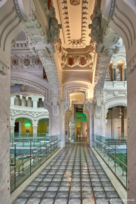 Palacio de Cibeles, Madrid Ornate interior of a grand building with an arched ceiling decorated with intricate carvings and emblems. The glass-tiled walkway is flanked by marble columns, leading towards a brightly lit doorway.