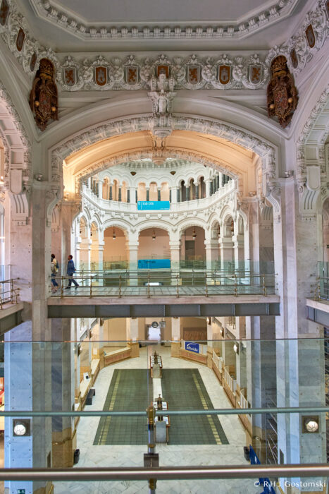 A grand, ornate interior inside Palacio de Cibeles features arched balconies, decorative carvings, and large glass balustrades. People walk on the upper level above a spacious ground floor adorned with marble accents.