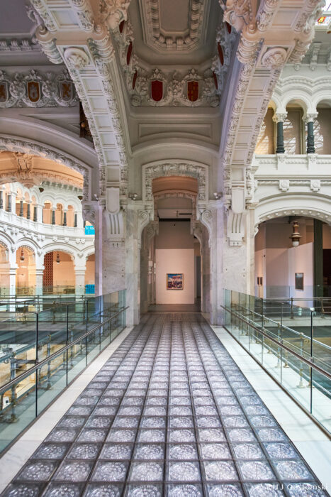 Palacio de Cibeles, Madrid A grand, ornate interior corridor inside Palacio de Cibeles, Madrid, features a patterned glass floor, arched doorways, detailed white vaulted ceilings, and decorative shields. Glass railings line the walkway leading to framed artwork on the far wall.