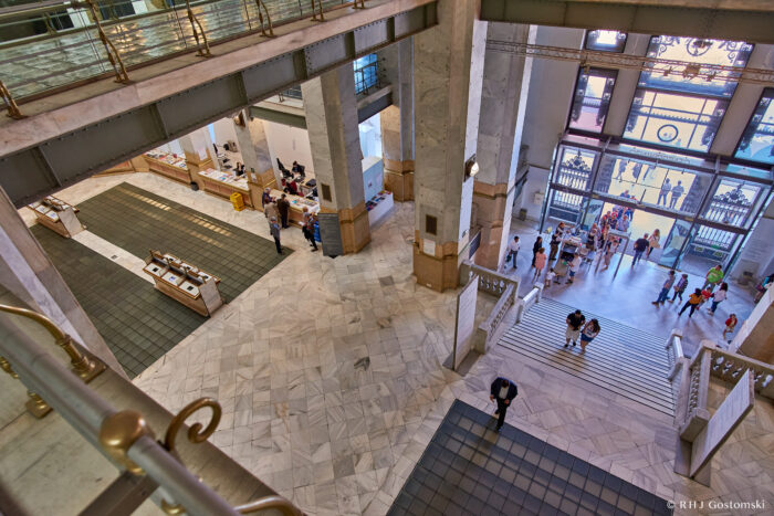 Interior view inside Palacio de Cibeles, showing a spacious marble-floored hall with people near stairs and counters on the left. Large windows and decorative columns fill the well-lit, elegant space.