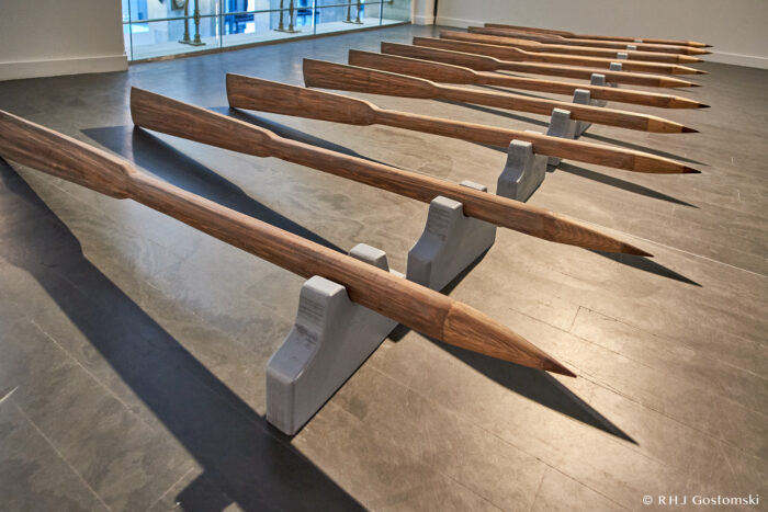 Palacio de Cibeles, Madrid Several large, wooden oars with pointed ends are arranged in parallel rows on a grey floor, each supported by grey stands in a brightly lit indoor space.