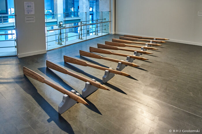 Rows of large, pointed wooden stakes are displayed horizontally on metal supports in a modern indoor gallery with grey flooring and glass railings, creating a striking art exhibit at CentroCentro.